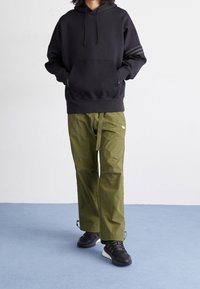 En person med svart hoodie, olivgröna byxor med bälte och svarta sneakers står på ett ljusblått golv mot en vit vägg.