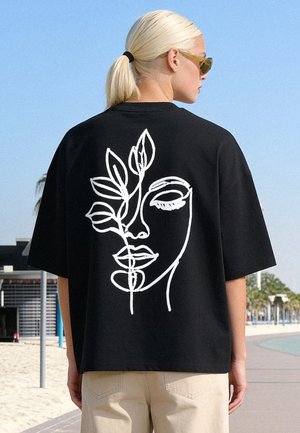 Personne aux cheveux blonds en queue de cheval, portant des lunettes de soleil et un t-shirt noir avec un motif abstrait blanc de visage et de feuille dans le dos, en extérieur.