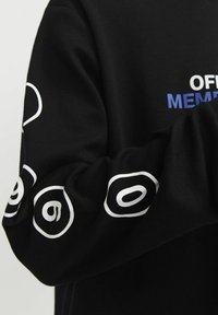 Camisa negra de manga larga con patrones gráficos blancos en las mangas y texto azul que dice "OFF MEM" en el pecho. Tejido de algodón suave.