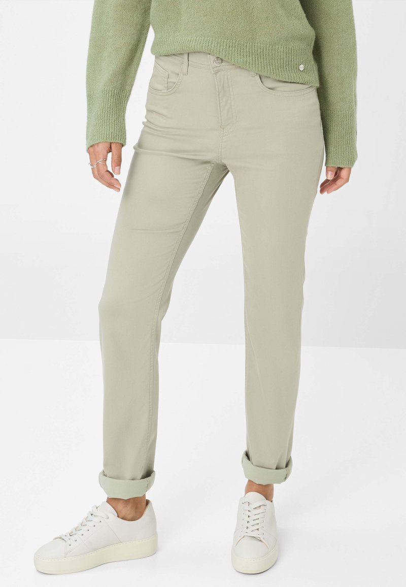 Chino en coton vert clair avec une coupe droite, ourlets roulés, poches avant et une texture lisse, associés à des baskets blanches.