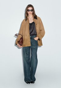 Tan suède jacket met een zwarte kraag, zwarte tanktop, blauwe wijde jeans en een bruine handtas. Model draagt zwarte zonnebril.