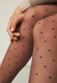 Transparante bruine panty met zwarte stippen, met een gladde textuur en een aansluitend ontwerp, gedeeltelijk zichtbaar op benen met een hand die rust.