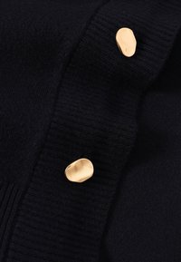 Pull en maille bleu marine avec texture côtelée et trois boutons dorés de forme irrégulière sur le devant pour la fermeture.