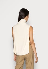 Bezrukávový hřejivý rolák v krémové barvě s otevřeným zadním dílem. V kombinaci s béžovými kalhotami, které mají měkkou texturu a volný střih.