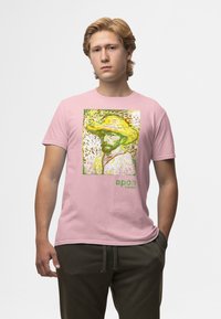 Camiseta de algodón rosa con un estampado gráfico de un hombre con un sombrero de paja y acentos verdes, etiquetada como "apoh London". Textura suave, diseño casual.