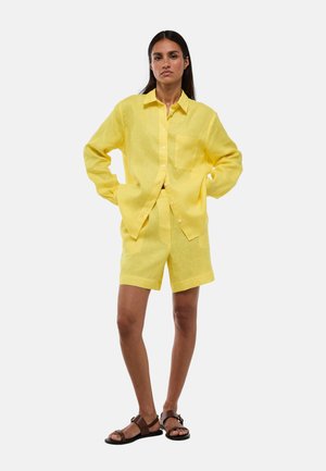 Femme debout, les mains dans les poches, portant une chemise en lin jaune à boutons et un short assorti avec des sandales marron sur fond blanc.