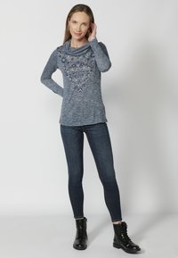 Modrá, dlhý rukáv, top s kaskádovým golierom a zložitou kvetinovou výšivkou. Nosený so slim fit tmavými džínsy a čiernymi šnurovacími topánkami.