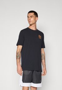 Sort bomulds t-shirt med korte ærmer og rund hals, prydet med orange grafisk tekst på venstre bryst. Båret med mesh shorts i sort og hvid.