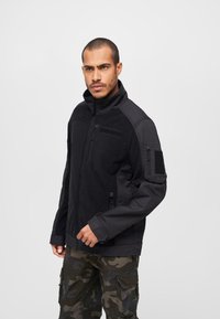 Black zip-up jacket featuring fleece material, high collar, and multiple pockets. Notable details include a Velcro patch on the left arm.