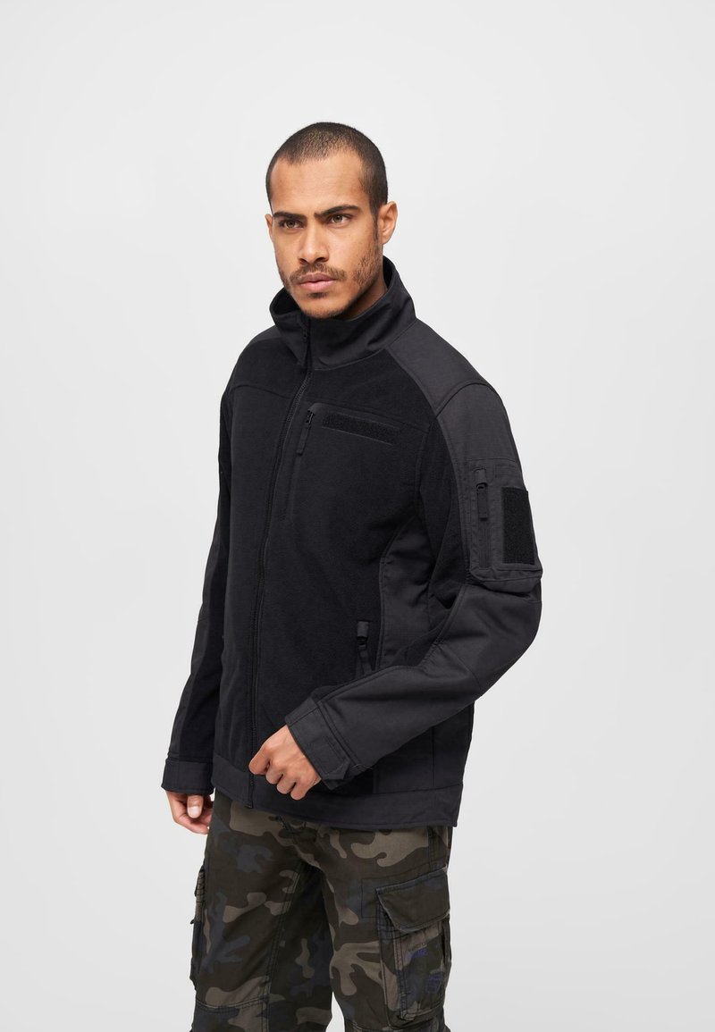 Black zip-up jacket featuring fleece material, high collar, and multiple pockets. Notable details include a Velcro patch on the left arm.