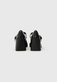 Black patent leather wedge heels with a textured, solid base and a pointed toe. Features a strap with a buckle for secure fastening.