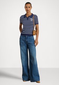 Námornícka modrá a biela prúžkovaná polokošeľa s golierom, doplnená širokými modrými džínsami. Malá hnedá kabelka nosená cez rameno.