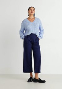 Hellblauer Strickpullover mit V-Ausschnitt, Rautenmuster und weiten Ärmeln, kombiniert mit dunkelblauen weit geschnittenen Hosen. Schwarze Slipper runden den Look ab.