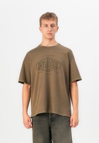 Ung mann iført en løs brun t-skjorte med "Dry Goods"-logo og mørke jeans, stående foran en ensfarget hvit bakgrunn.