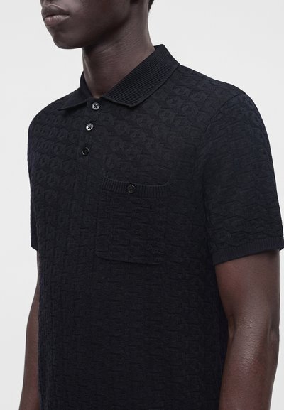 Homme portant un polo noir texturé à manches courtes avec trois boutons et une poche poitrine boutonnée sur un fond uni.