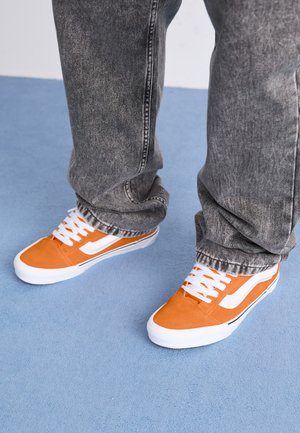 Oransje semskede sneakers med hvite striper og snørebånd, kombinert med falmede grå jeans. Plassert på et lys blått teppe.
