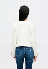 Sweater branco, texturizado, com um design cropped, decote redondo e detalhes em botão nos punhos, combinado com jeans azuis com bolsos traseiros.