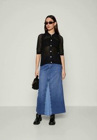 Ženska v črni mrežasti bluzici s gumbi, dolgem modrem jeans krilu s sprednjo špranjo, črnih čevljih, sončnih očalih, drži majhno črno torbico.