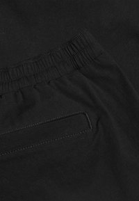 Nahaufnahme von schwarzen Stoffhosen mit elastischem Bund und einer abgesteppten, horizontalen Gesäßtasche.