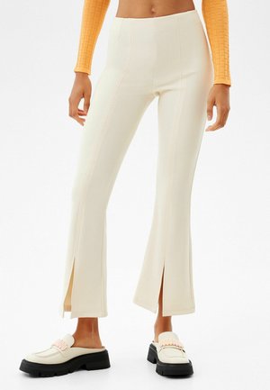 Femme portant un pantalon crème taille haute avec des fentes sur le devant des jambes, associé à des mocassins crème épais et un top orange court à manches longues.