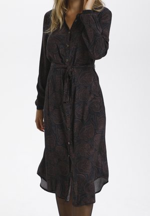 Vrouw draagt een midi-jurk met lange mouwen en knopen, met een donker paisley patroon, taille gestrikt, staand tegen een egale lichte achtergrond.