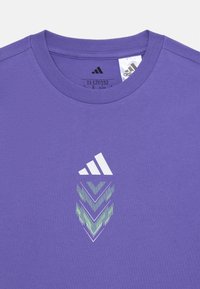 Lilla bomull t-skjorte med rund hals. Den har en hvit adidas-logo og et geometrisk design i grønn og hvit under logoen.