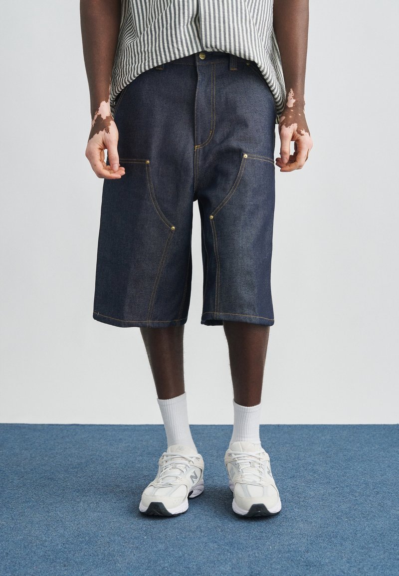 Άτομο που φορά ριγέ πουκάμισο, μακριές σκούρες τζιν βερμούδες, λευκές κάλτσες και λευκά αθλητικά παπούτσια, στέκεται σε μπλε χαλί με ορατό λεύκη στα χέρια.