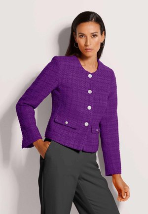 Vrouw draagt een paarse gestructureerde jas met zilveren knopen en zwarte broek, poseert met één hand in de zak tegen een effen achtergrond.