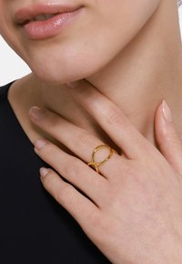 Goldene Ring mit einem ineinander verwobenen Design, auf einer Hand mit natürlichen Nägeln präsentiert. Hautton und Ring betonen den minimalistischen Stil.
