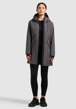 Frau steht vor einem schlichten hellgrauen Hintergrund, trägt einen grauen Kapuzenmantel, schwarze Leggings und schwarze Turnschuhe.