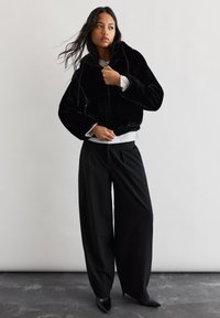 Črna dukserica iz umetnega krzna s kapuco in vrvico, v kombinaciji s širokimi črnimi hlačami. Model nosi belo majico pod dukserico.