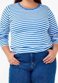 Blå och vitrandig långärmad topp med rundad halsringning, tillverkad av ribbat material. Tillsammans med mörkblå jeans, händerna placerade på höfterna.