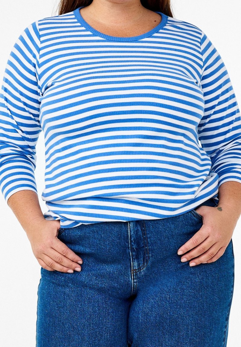 Blå och vitrandig långärmad topp med rundad halsringning, tillverkad av ribbat material. Tillsammans med mörkblå jeans, händerna placerade på höfterna.