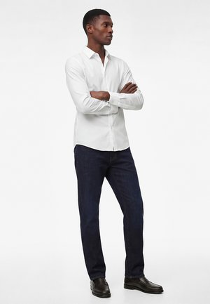 Mann steht mit verschränkten Armen, trägt ein weißes Hemd mit Knöpfen, dunkelblaue Jeans und schwarze Anzugschuhe vor einem weißen Hintergrund.