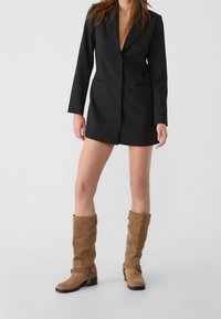 Blazer nero su misura con rever a punta, dalla vestibilità slim, abbinato a stivali al ginocchio in suede color cammello con dettagli in cinturino e fibbia.