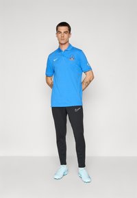 Mand står med hænderne bagved ryggen, iført en lys blå polo med logo, sorte Nike-bukser og lyseblå fodboldstøvler på en ensfarvet baggrund.