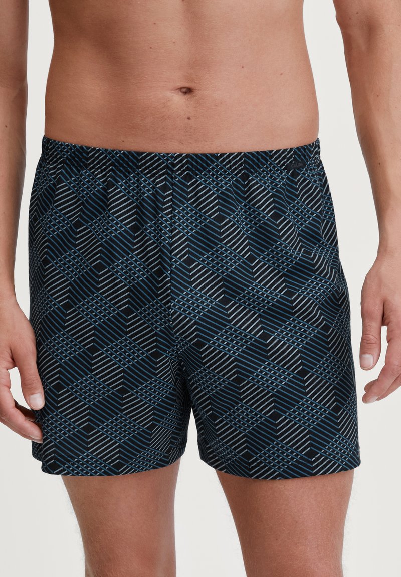 Zwarte zwemshorts met een geometrisch patroon in blauw en wit. Elastische tailleband zonder zichtbare hardware. Gladde stoftextuur.