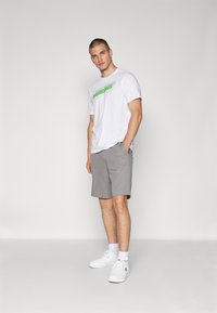 Camiseta blanca de algodón con un logo verde, combinada con pantalones cortos grises de tejido suave. El modelo lleva zapatillas blancas y calcetines blancos.