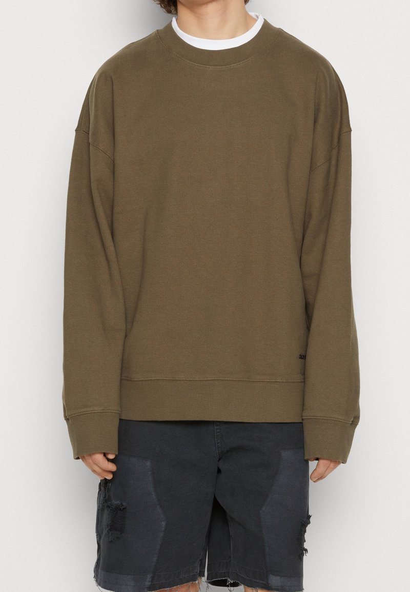 Personne portant un sweat-shirt vert olive oversize par-dessus un t-shirt blanc et un short en jean noir déchiré.