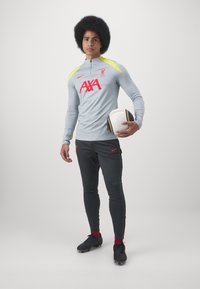 Camiseta de entrenamiento gris de manga larga con detalles amarillos y un logo rojo, combinada con pantalones negros ajustados y botas negras. El jugador sostiene un balón de fútbol blanco.
