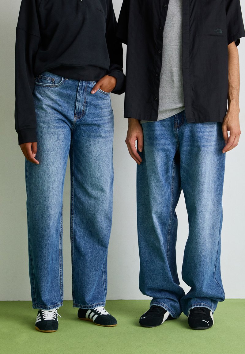 To personer iført lyseblå, løstsittende denimjeans. En person har på seg hettegenser i svart, den andre en grå skjorte og svart jakke.