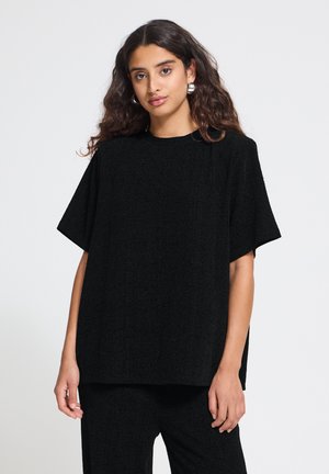 Modèle portant un haut noir ample à manches courtes et un pantalon assorti, avec des boucles d’oreilles argentées, sur un fond clair uni.