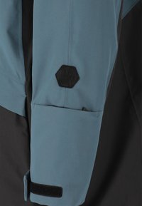 Blaue und schwarze Jacke mit schlankem Design, glattem Stoff, verstellbarem Bündchen mit Klettverschluss und einem Sechseck-Logo auf dem Ärmel.