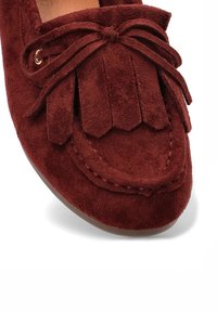 Mocasín de ante marrón con solapa decorativa de flecos y lazo atado en la parte delantera, con detalles de costura a lo largo de la zona del dedo del pie.