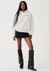 Sweat-shirt blanc avec le texte "BLACKPINK" rose, coupe oversize, poche kangourou. Associé à une mini-jupe noire et des bottes noires brillantes jusqu'au genou.
