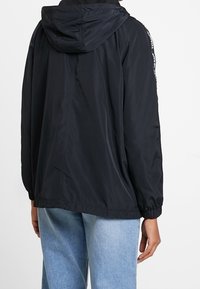Černá lehká bunda s kapucí, elastickými manžetami a lemováním, se znakem na rukávech. Hladká textura a volný střih.