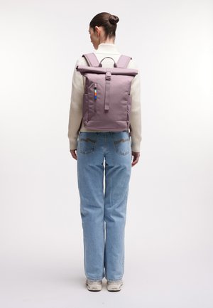 Woman with hair in bun wearing a beige sweater, light blue jeans, white sneakers, and a mauve roll-top backpack facing away on white background.