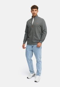 Hombre vestido con chaqueta gris de cremallera, jeans azul claro y zapatillas blancas, de pie contra un fondo blanco.