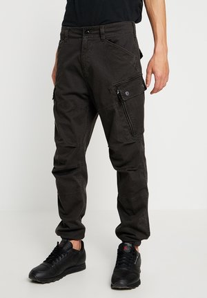 Pantalon cargo noir avec plusieurs poches, porté avec des baskets noires et une chemise noire, montré de la taille aux pieds.