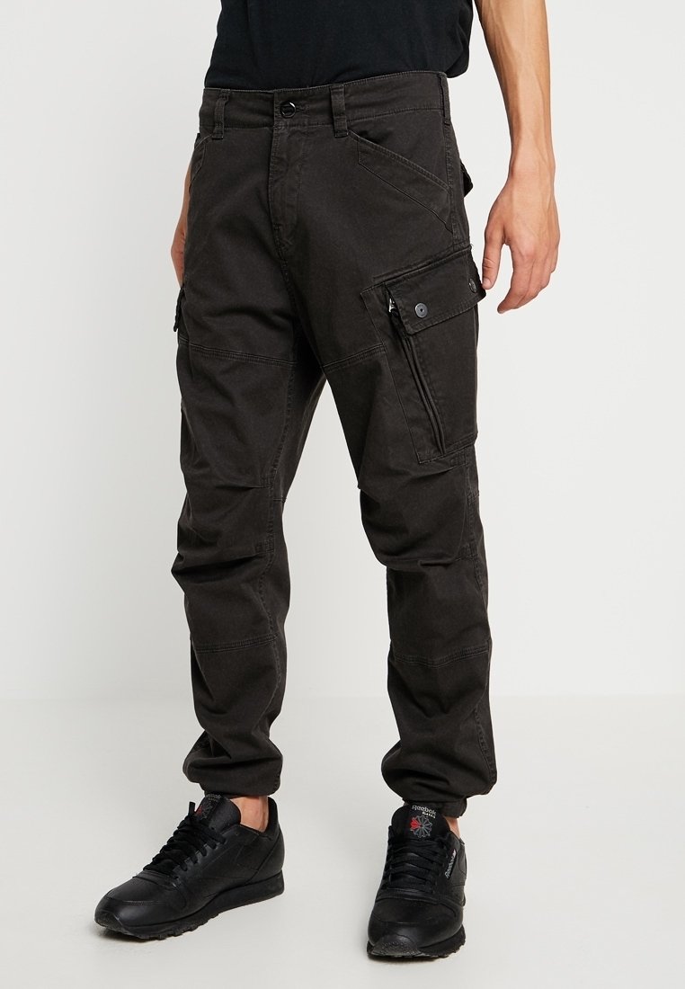 Pantalon cargo noir avec plusieurs poches, porté avec des baskets noires et une chemise noire, montré de la taille aux pieds.